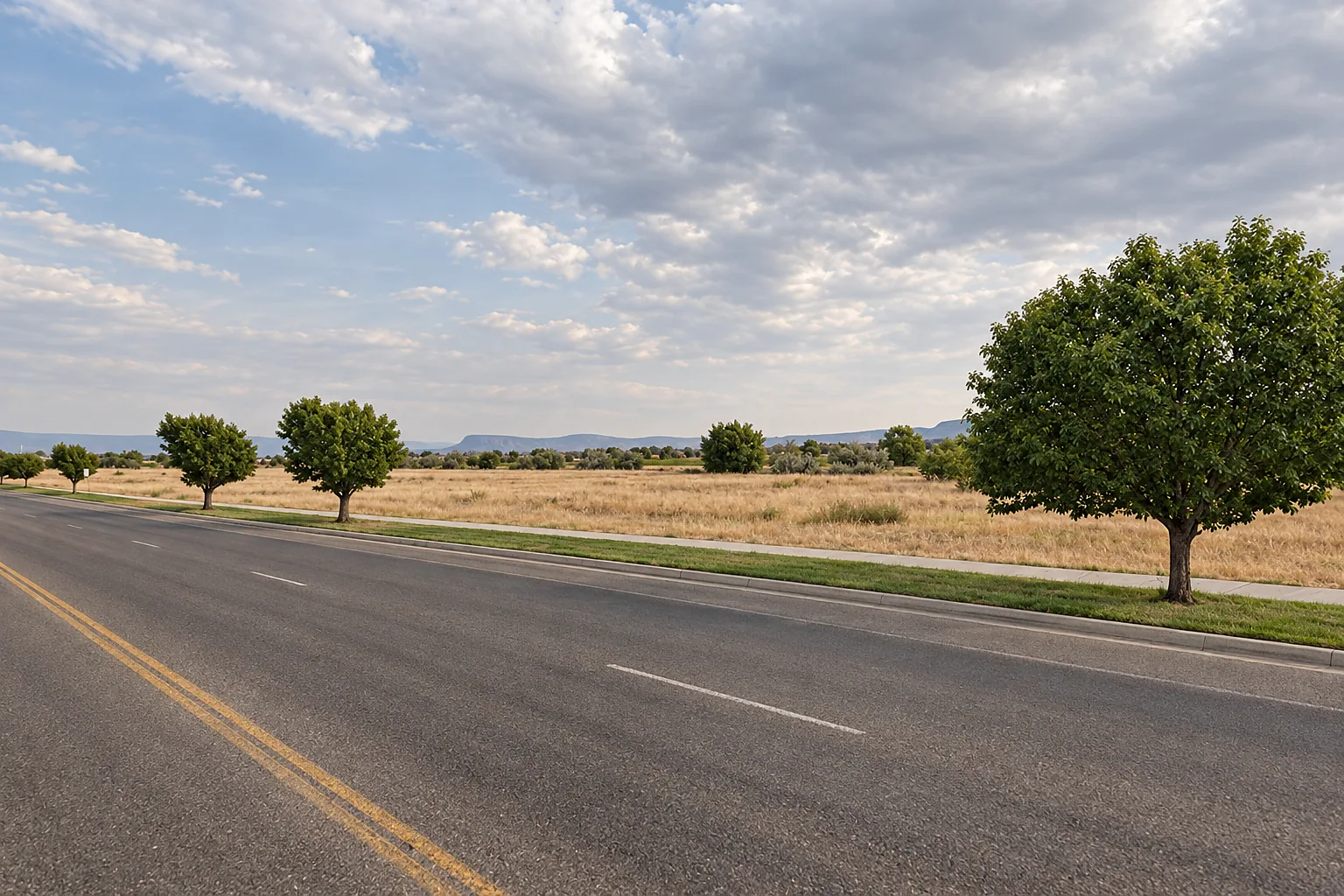 Open rangeland beside a wide main road west of Vernal, with roadside trees and mountains on the horizon